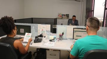 atendentens de costas para foto estão diante de computadores. A mulher, à esquerda, segura um folheto do cadoj. Ao fundo há um homem sentado a uma mesa.#paratodosverem 