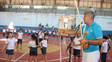 homem toca berimbau e canta próximo a microfone. Crianças estão na quadra da escola. #paratodosverem 