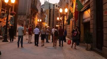 pessoas circulam em calçadão de rua ao cair da tarde. Luminárias estão acesas. #paratodosverem