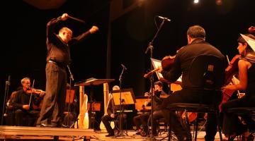 maestro em pé rege orquestra em palco. #paratodosverem