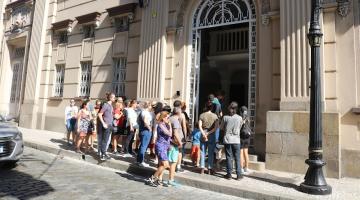 pessoas estão paradas em frente a prédio histórico no centro, e ao lado de poste tipo republicano. #paratodosverem 