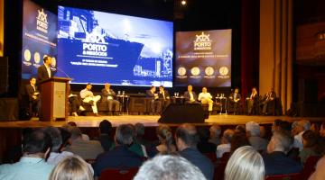 Palco de teatro com autoridades sentadas e um homem falando em uma tribuna. Em primeiro plano está a plateia, de costas para a imagem. #Pracegover