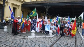 várias pessoas com trajes típicos e bandeiras de seus países posam para foto diante da estação do valongo. #paratodosverem