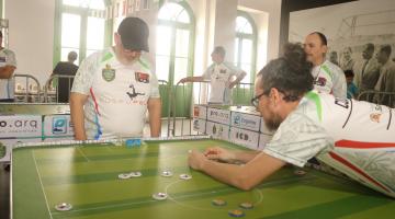 dois homens jogam botão em salão. #paratodosverem