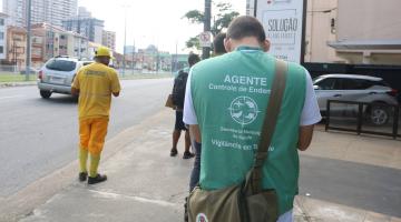agente de combate à dengue caminha em rua, de costas para a foto, com evidência no colete verde. Ao lado esquerdo, um funcionário da terracom. #paratodosverem 
