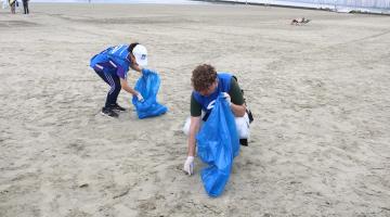 duas pessoas estão na faixa de areia coletando resíduos e levando-os a um saco plástico. #paratodosverem 