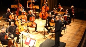 O grupo sinfônico de Santos em exibição em teatro. Os músicos estão dispostos no palco com seus instrumentos e diante de suas estantes com partituras. O maestro está à frente de todos sobre um púlpito. #Pracegover