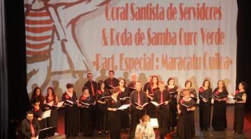 Coral está em palco. Ao fundo há um painel onde se lê Coral Santis de Servidores & Escola de Samba Ouro Verde Part.Especial: Maracatu Quiloa. #Pracegover