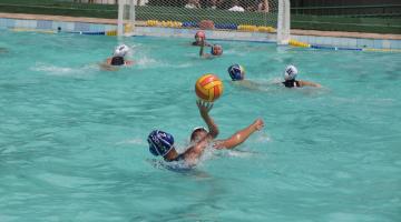 A piscina com atletas jogando polo aquático. Ao fundo está a trave. #Pracegover