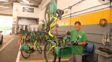 Homem conserta bicicleta. há bikes ao fundo do galpão. #paratodosverem