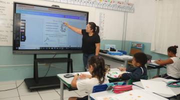 sala de aula com professora em pé falando aos alunos. #paratodosverem