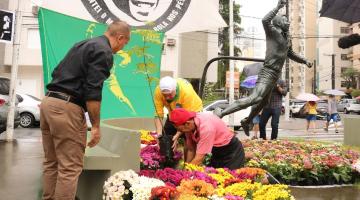 dois homens enterram muda de árvore no solo junto a florres. Ao fundo, uma bandeira com a imagem de pelé, e ao lado direito, a estátua do Rei. #paratodosverem