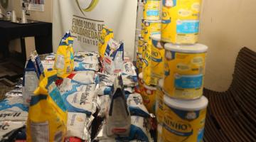 latas e pacotes de leite em pó sobre uma mesa, com banner do fundo social ao fundo. #paratodosverem