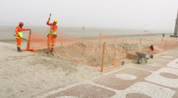 dois homens estão cercando com lona plástica perfurada trecho de areia na praia, junto ao calçadão. #paratodosverem 
