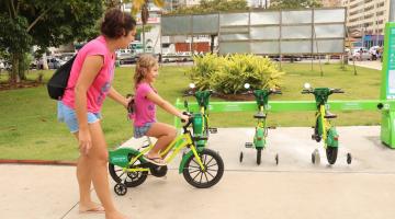criança pedala bicicleta com a mãe em pé ao lado. #paratodosverem