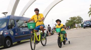 homem adulto e criança andam de bicicletas em parque. Ao lado deles está parada uma viatura da guarda municipal. #paratodosverem 