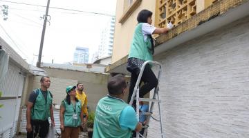 mulher usa escada para visualizar calha em prédio. Ela está sendo observada por outros agentes de combate à dengue. #paratodosverem 