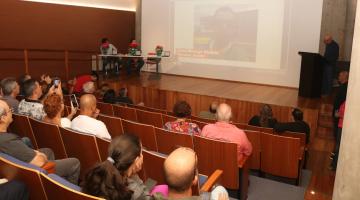 pessoas sentadas em auditório assistindo palestra e a imagens num telão ao fundo. #paratodosverem 