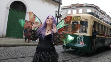 mulher com longo cabelo roxo usa asas de borboleta. O bonde está atrás dela. #paratodosverem 