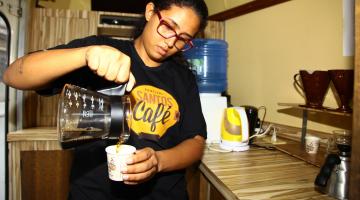Mulher serve café. Ela vira o conteúdo da bebida em uma jarra em um copo. Em sua camiseta está estampado o logotipo do festival. #Pracegover