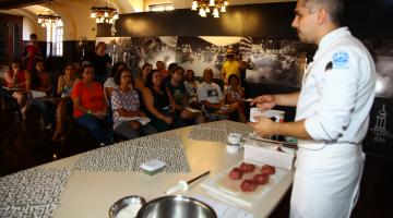 Aula de culinária dentro de sala de museu. O chefe está à frente de mesa, diante de uma tábua com alimentos. À frente está o público sentado assistindo a aula. #Pracegover