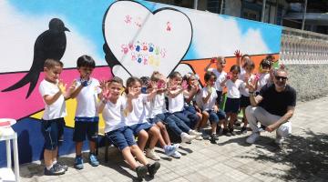 crianças estão sentadas em banco diante de arte pintada em muro. Elas esticam as mãos para a frente posando para a foto. #paratodosverem 