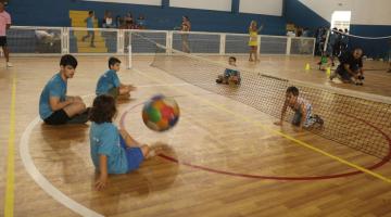 crianças jogam bolam em quadra. Todas estão sentadas. Há uma rede dividindo a quadra para o vôlei sentado. #paratodosverem 