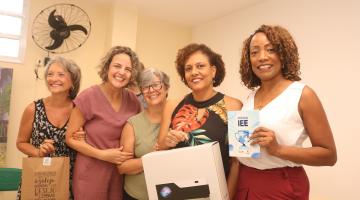 mulheres seguram pacote com projetor e azulejo decorado. #paratodosverem