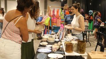 estande de venda de artesanato com duas consumidoras olhando os produtos e uma mulher, do outro lado da mesa, atendendo. #paratodosverem 