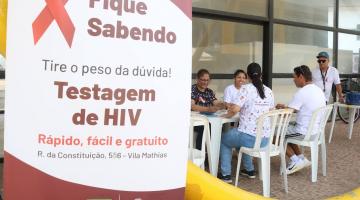 banner onde se lê Fique sabendo tire o peso da dúvida testagem de hiv. Ao fundo, pessoas fazem exames. #paratodosverem 