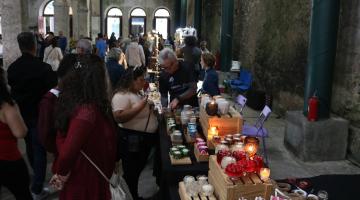 estande com exposição de velas em primeiro plano e pessoas circulando em um salão. #paratodosverem 