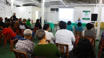 Salão com público sentado às cadeiras. As pessoas estão atentas a um homem que fala ao grupo. Ele está ao fundo diante de um painel. #Pracegover