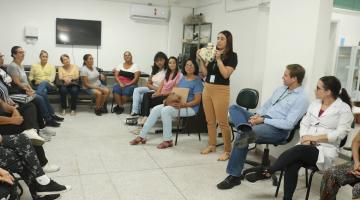 pessoas estão sentadas em círculo assistindo palestra. uma mulher fala em pé ao grupo. #paratodosverem