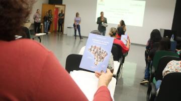 pessoas assistindo palestra. Em primeiro plano, uma mulher, de costas, segura um folheto com instruções para o cuidado com idosos. #paraatodosverem 