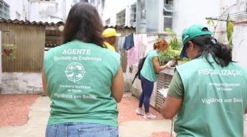 duas agentes da dengue uniformizadas, de costas para a foto. Ao fundo uma agente vistoria vaso em área aberta de uma casa. #paratodosverem 