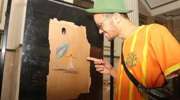 artista aponta para uma de suas obras afixada em display. #paratodosverem 