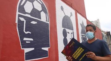 Artista segura livro em frente de parede com colagens #paratodosverem