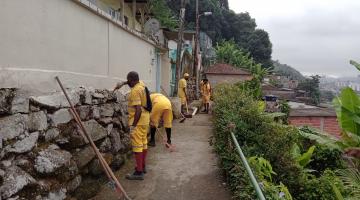 homens uniformizados raspam pedras em muro de contenção em morro. Ao lado direito há vegetação. #paratodosverem