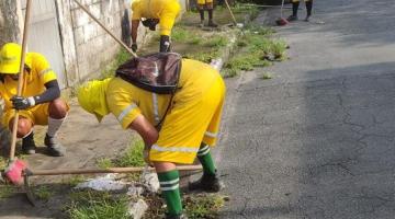 homens uniformizados raspam calçada cheia de mato. #paratodosverem