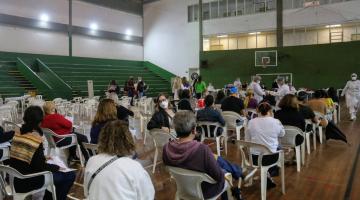 Pessoas estão sentadas em cadeiras afastadas em ginásio esportivo. #paratodosverem