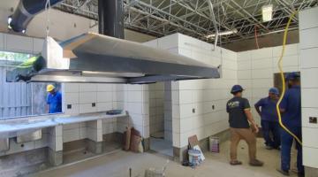 homens trabalham em área que é uma cozinha. Há obras no local. É possível ver a estrutura de uma coifa industrial. #paratodosverem 