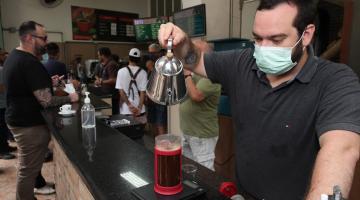 HOMEM SEGURA BULE E DESPEJA ÁGUA EM COPO PREPARANDO UMA BEBIDA DE CAFÉ NUM COPO LONGO. MOVIMENTAÇÃO DE PESSOAS AO FUNDO. #PARATODOSVEREM