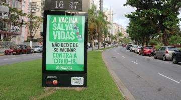 toten com relógio informando a hora no topo. Abaixo, orientação sobre vacina. Toten está em canteiro central na avenida da oral. Os prédios estão do lado esquerdo. A pista à direita está sem movimento, somente com veículos estacionados. #paratodosverem