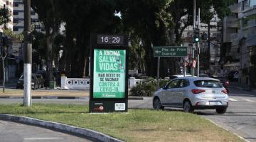 Totem em canteiro central com horário marcando 16h29. O restante do totem tem informação sobre a importância da vacina. Há um carro fazendo curva ao lado. #paratodosverem