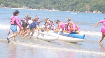 mulheres entram no mar, ainda empurrando a canoa havaiana. Morro ao fundo. #paratodosverem