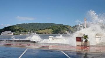 Em ressaca, ondas do mar batem na mureta da ponta da praia. #paratodosverem