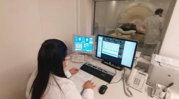 Mulher está sentada em sala diante de computador. Ao fundo em outra sala, é realizada uma ressonância. #Paratodosverem
