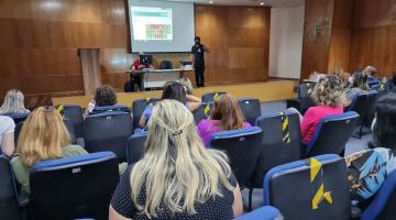 Pessoas sentadas em auditório assistindo palestrante #paratodosverem