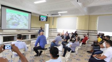 pessoas estão sentadas em cadeiras distribuídas em salão. Todas assistem a uma projeção num telão e estão atentas a um homem que fala à frente de todos.#paratodosverem