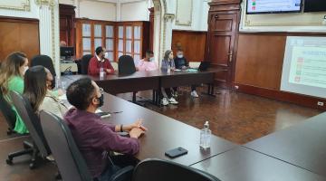 pessoas sentadas em torno de mesa em formato de U. Elas olham para um telão onde há informações escritas. #paratodosverem